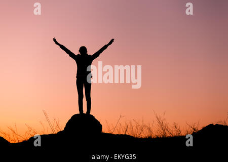 Silhouette of woman with arms raised up contre beau ciel coloré. Coucher du soleil d'été. Paysage Banque D'Images