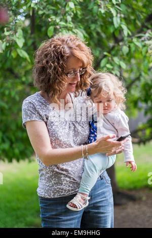 Une grand-mère âgée de 50 ans tient son enfant petite-fille sur sa hanche tout en plaçant un appel mobile à l'extérieur. Banque D'Images