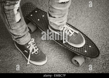 Jeune skateur professionnel en jeans et gumshoes debout sur son skate. Close-up fragment de skateboard et pieds, stylets rétro monochrome Banque D'Images