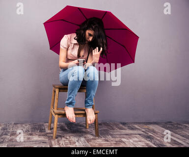 Jeune femme assise sur la chaise avec le smartphone et un parapluie rose. En portant des jeans et chemise. Banque D'Images
