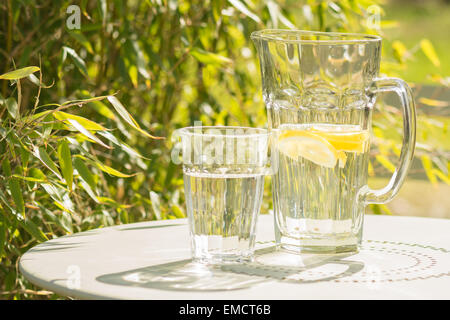 L'été - Reflets de soleil qui brille à travers le verre et carafe d'eau de citron vert sur la table de bistro sur terrasse de jardin Banque D'Images