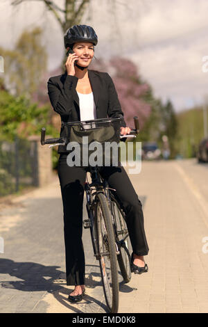 Cet élégant young businesswoman riding au travail s'arrêter pour répondre à un appel sur son téléphone portable à l'appui permanent de son vélo Banque D'Images