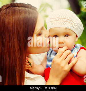 Surpris Toddler Baby Girl sitting on Mom's Hands, à la recherche en appareil photo. Mère embrassant l'enfant. Détente dans le parc d'été vert, Banque D'Images