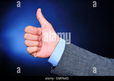 Libre de la main d'un jeune homme de race blanche portant un costume gris donnant une thumbs-up signe sur un fond noir allégé Banque D'Images