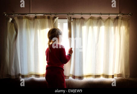 Une petite jeune fille en pyjama ouvre les rideaux pour regarder par la fenêtre au début d'un nouveau jour Banque D'Images