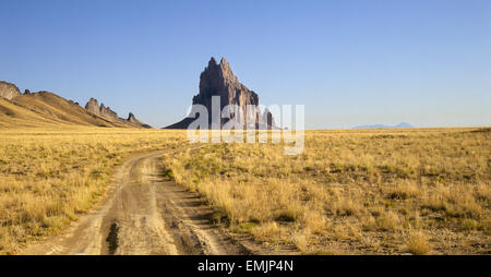 Rutty un chemin de terre mène à Shiprock, un pic volcanique sur la réserve indienne Navajo dans le nord du Nouveau Mexique. Aussi appelé le Rock avec des ailes par les Navajos, le pic est considéré comme sacré. Dans l'arrière-plan est en train de dormir Ute Mountain, considérée comme sacrée par les Indiens du SEI. Banque D'Images