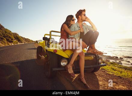 Ambiance jeune couple assis sur le capot de leur voiture le long d'une route côtière ouverte avec l'homme répondre à un appel téléphonique et d'une femme si Banque D'Images