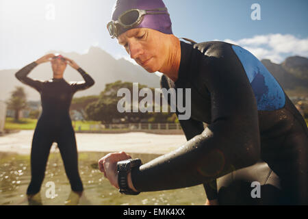 Jeune homme concentré contrôler sa montre alors qu'en combinaison de plongée au bord du lac. Man looking at watch après échauffement. Les triathlètes. Banque D'Images