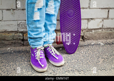 Adolescent en jeans et gumshoes stands avec près de skateboard urbain mur gris Banque D'Images