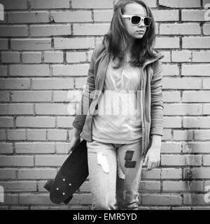 Adolescente blonde en jeans et lunettes détient plus de skateboard urban mur de briques, l'arrière-plan photo monochrome Banque D'Images