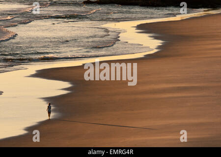 Figure solitaire promenades au bord de l'eau sur la plage au lever du soleil, le début de l'incandescence du matin jette une ombre sur le sable. Banque D'Images