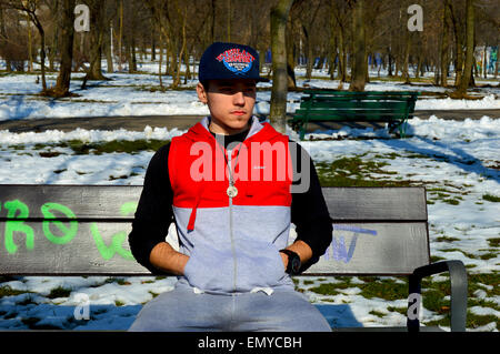 Man wearing hoodie, cap et collier assis sur banc de parc Banque D'Images