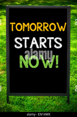 Demain commence maintenant ! Message sur trottoir tableau noir signer contre fond d'herbe verte. Copie de l'espace disponible. Image Concept Banque D'Images
