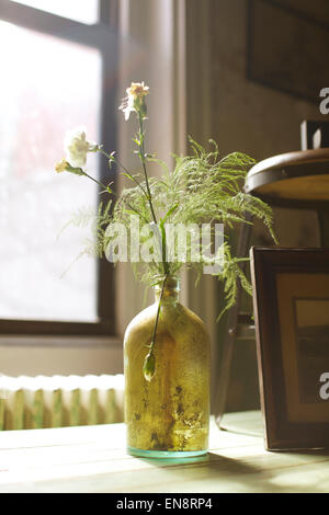 Une bouteille vintage sale habilement utilisé comme un vase à fleurs d'oeillets blancs se trouve sur une surface en bois devant une fenêtre. Banque D'Images