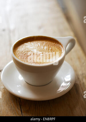 Un cappuccino avec un motif de feuilles dans la mousse dans une tasse blanche avec une soucoupe assis sur une table en bois, l'arrière-plan est flou. Banque D'Images