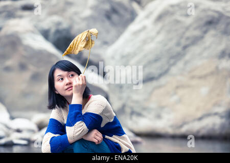 Young Asian woman sitting près d'un étang à un désert de pierre avec une grande feuille jaune dans sa main Banque D'Images