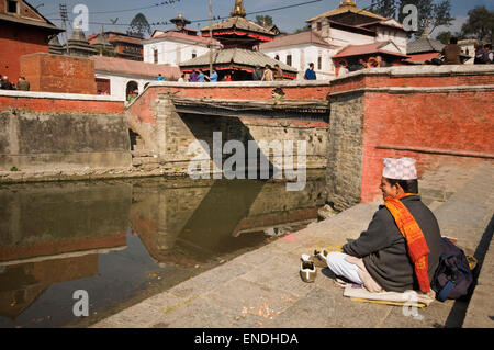 Le Népal, Katmandou, Pashupatinath, l'homme en costume traditionnel assis à côté de la rivière Bagmati (affluent du Gange) avec Pas Banque D'Images