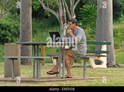 L'homme assis à une table de pique-nique dans un parc public à l'aide d'un ordinateur portable - Fond d'arbres et végétation verte émeraude Banque D'Images