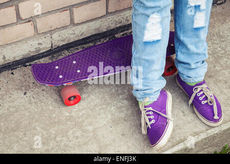 Adolescent en jeans et gumshoes stands avec près de skateboard urbain gris, fragment de mur pieds Banque D'Images