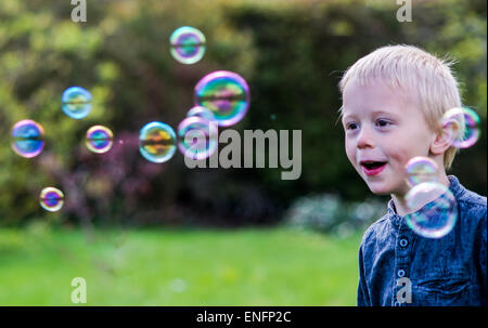 Un petit garçon souffle des bulles de savon dans le jardin un jour d'été. Il porte une chemise bleue et il est heureux Banque D'Images