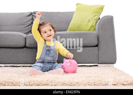Portrait of a cute little girl mettant l'argent dans une tirelire assis sur le sol en face d'un canapé gris moderne Banque D'Images