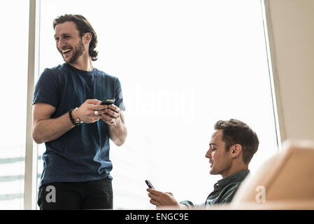 Deux hommes dans un bureau, la vérification de leurs téléphones intelligents. L'un à la suite de rire. Banque D'Images