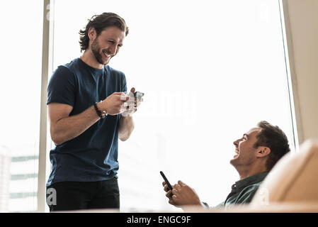 Deux hommes dans un bureau, la vérification de leurs téléphones intelligents. L'un regardant en riant. Banque D'Images