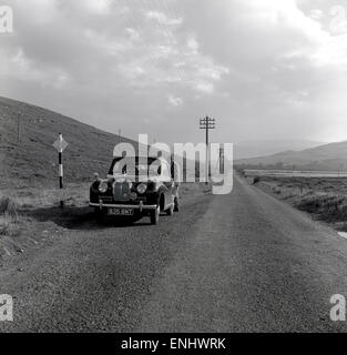 Années 1950, historique, un homme à l'arrière de sa voiture Austin Morris a garé sur un côté d'une longue route de tarmac, droite et vide dans les Highlands écossais, en Écosse, au Royaume-Uni. Banque D'Images