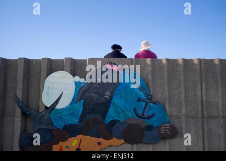 Vieux couple wearing hats assis par mer mur à signer le message écologique, Normandie, France Banque D'Images