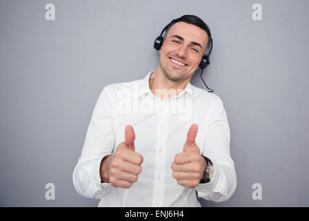 Heureux homme opérateur dans les écouteurs showing Thumbs up sur fond gris. Looking at camera Banque D'Images