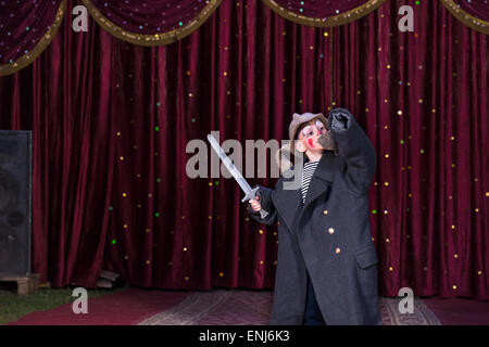 Young Boy Wearing Clown Maquillage et de grandes armoiries Holding Toy Prop Sword avec bras levé debout sur scène avec rideau rouge Banque D'Images