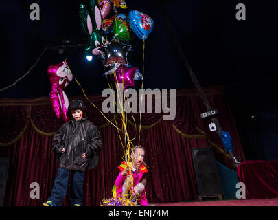 Jeune garçon habillé en clown portant veste en cuir et casque debout sur scène avec fille en costume de couleur dynamique Holding Ball Banque D'Images