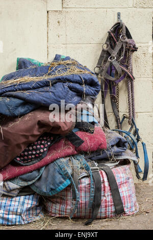 Pile de tapis et couvertures de cheval couverts dans le foin avec les chevaux chef des cols et entraîner des cordes de raccrocher dans une écurie. Banque D'Images