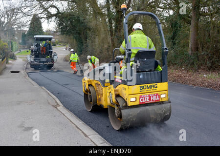 Les hommes qui travaillent sur la route font surface et répare les réparations à l'aide d'une machine d'épandage à rouleaux vibrants et à tarmac, portant des vestes et des chapeaux haute visibilité Essex UK Banque D'Images