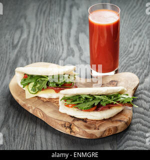 Sandwichs jambon rustique avec la roquette et les tomates dans le pain pita et verre de jus, sur table en bois Banque D'Images