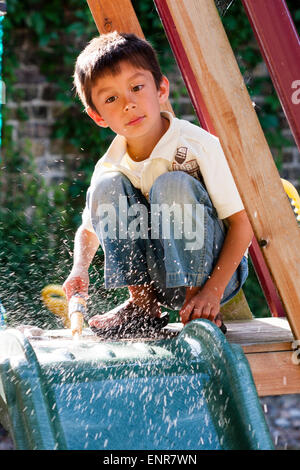 Race mixte, caucasien-asiatique enfant, garçon, 7-8 ans, agenouillé sur le dessus de la lame couverte, pulvérisant la lame nettoyer avec un tuyau à l'extérieur dans le jardin. Banque D'Images