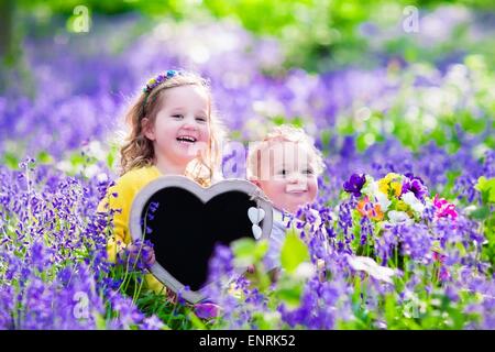 Enfants jouant dans bluebell flowers. Petite fille et garçon bébé tenir en forme de coeur en bois tableau. Copie de l'espace pour votre texte. Banque D'Images