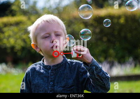 Un petit garçon souffle des bulles de savon dans le jardin un jour d'été. Il porte une chemise bleue et il est heureux Banque D'Images