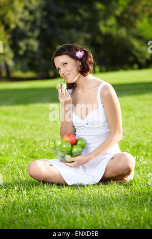Belle jeune femme est assise et mange une pomme sur la pelouse au jardin d'été Banque D'Images