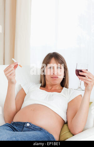 Belle femme enceinte tenant une cigarette et un verre de vin rouge en position allongée sur un canapé Banque D'Images