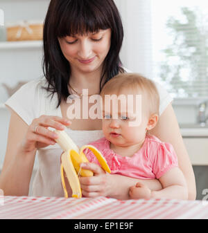 Charmante brunette woman peler une banane tout en tenant son bébé sur ses genoux dans la cuisine Banque D'Images