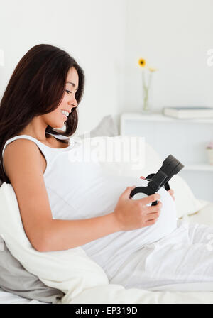 Charmante femme enceinte mettant le casque sur son ventre tout en étant allongé sur un lit Banque D'Images