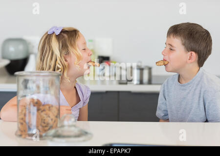 Frères et sœurs avec les cookies dans la bouche au comptoir de la cuisine Banque D'Images