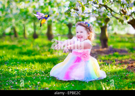 Enfant jouant avec un oiseau. Heureux rire petite fille en costume de fée avec des ailes de nourrir un perroquet dans un cerisier jardin Banque D'Images