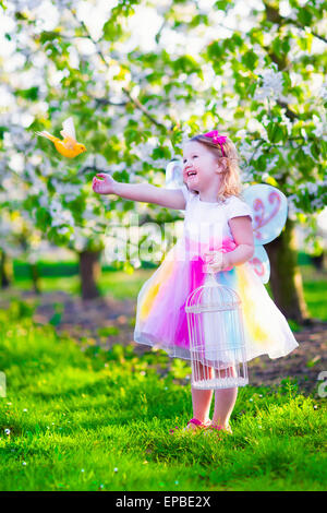 Enfant jouant avec un oiseau. Heureux rire petite fille en costume de fée avec des ailes de nourrir un perroquet dans un cerisier jardin Banque D'Images