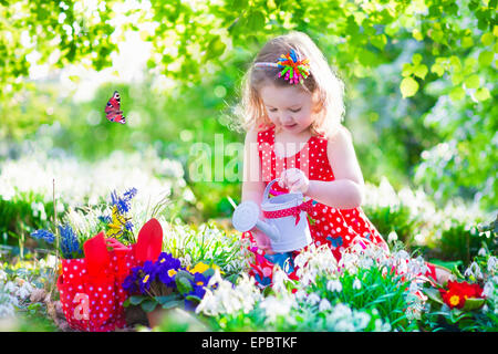 Cute little girl bouclés dans une robe d'été rouge travaillant dans le premier arrosage de jardin de fleurs de printemps sur une chaude journée ensoleillée Banque D'Images