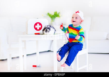 Cute laughing baby boy playing animal doctor avec son animal à l'aide de lapin toy stéthoscope dans une pépinière blanc prix Banque D'Images