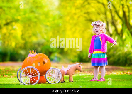 Cute little girl playing bouclés conte Cendrillon tenant une baguette magique à côté d'un chariot d'avoir citrouille in autumn park Banque D'Images