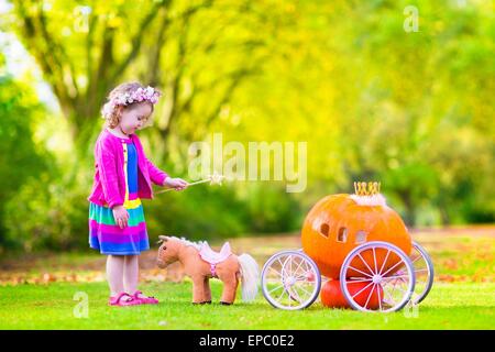 Cute little girl playing bouclés conte Cendrillon tenant une baguette magique à côté d'un chariot d'avoir citrouille in autumn park Banque D'Images