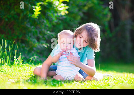 Deux frères, heureux petit garçon de l'école et un petit bébé, jouer ensemble assis sur la pelouse qui sent les fleurs Daisy Banque D'Images